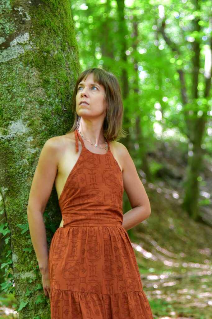 Femme debout adossée à un arbre dans la forêt, les yeux rivés au ciel, en pleine contemplation.